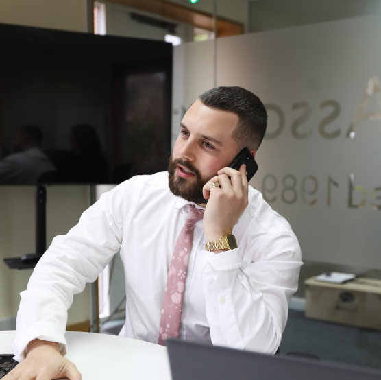 A man in a white shirt and pink tie talks on the phone while sitting at a desk in an office.