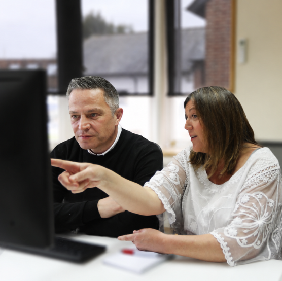 A man and woman sit together at a desk in a modern office, focused on a computer screen. The woman, wearing a white embroidered blouse, points toward the monitor while speaking, and the man in a black sweater listens attentively. Large windows in the background let in soft natural light, and desktop monitors frame the scene on both sides.